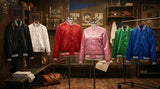 Six True Varsity satin bomber jackets displayed in a vintage locker room setting in black, white, red, pink, green, and blue with ribbed stripe trims and snap button fronts.