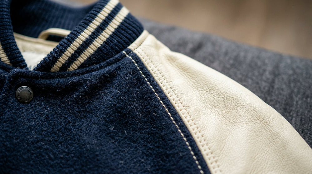 Extreme close-up of a navy wool varsity jacket featuring cream leather raglan sleeves and detailed double-needle stitching.